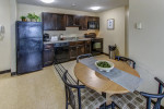 Modern kitchen with dark cabinets, dishwasher, refrigerator, and dining area in a furnished apartment.