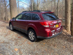 2011 Subaru Outback in red, parked on gravel surrounded by trees, showing a scuff on the rear bumper.